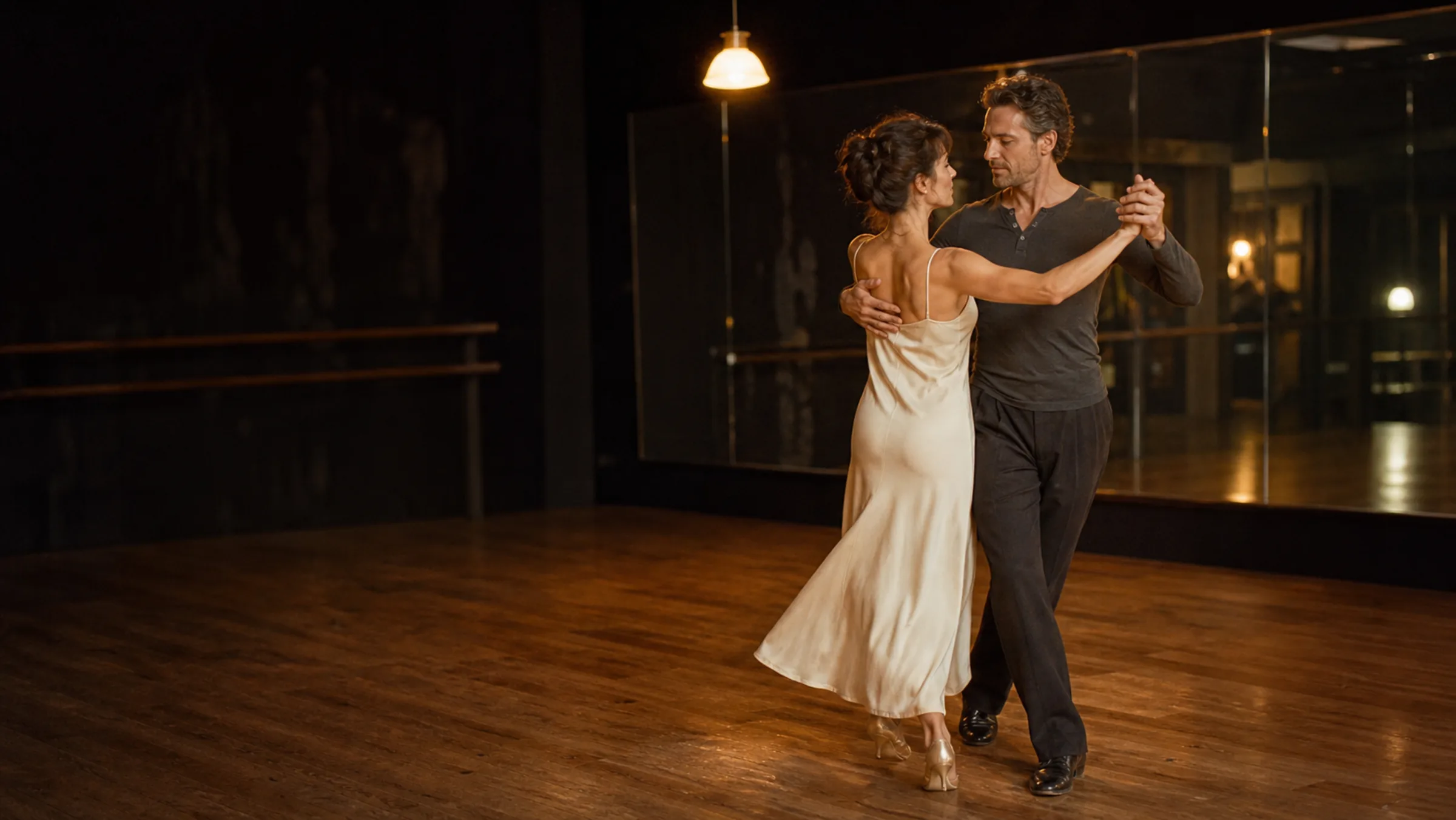 A couple in a slow ballroom hold on a warm wood floor under a single tungsten lamp at S-Style Dance Team